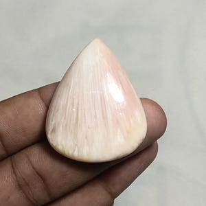 Può includere: Una gemma lucida a forma di goccia con una superficie liscia e brillante, caratterizzata da un motivo fibroso radiale color rosa pallido e crema che si irradia dalla base verso la punta, mettendo in risalto riflessi di luce naturali.