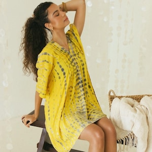 Puede incluir: Un vestido amarillo brillante hasta la rodilla con escote en V, mangas cortas y un patrón vertical de teñido anudado en tonos grises. La prenda tiene una silueta holgada y fluida con detalles plisados en el escote y un dobladillo recto.
