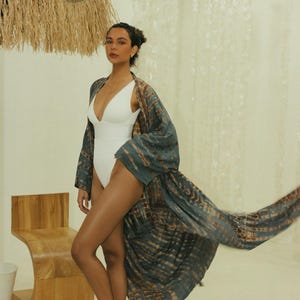 May include: A white one-piece swimsuit with a deep V-neckline paired with a long, flowing kimono robe featuring a blue and brown tie-dye pattern. The robe has wide sleeves and an open front, draping loosely over the white bodysuit.