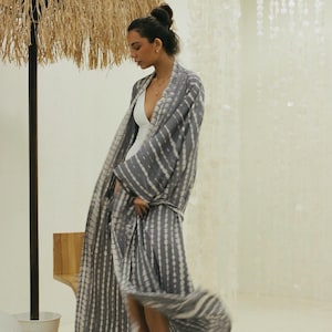 Puede incluir: Un kimono largo abierto con un patrón tie-dye gris y blanco de rayas verticales. La prenda tiene mangas anchas y una silueta fluida, usada sobre un body blanco sencillo con un escote en V profundo y tirantes finos.