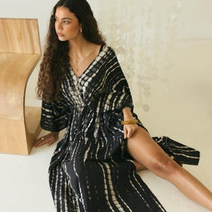 May include: A floor-length black and white tie-dye kaftan dress featuring a deep V-neckline, wide sleeves, and a cinched waist with a high side slit. The fabric displays a vertical pattern of dark black sections contrasted by white stripes.