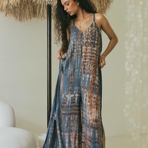 Op de afbeelding: Een mouwloze jumpsuit met een blauw en bruin tie-dye patroon, voorzien van een V-hals, dunne schouderbandjes en wijde broekspijpen. Het kledingstuk heeft een ontspannen, vloeiend silhouet met een verticaal kleurverloop in de stof.