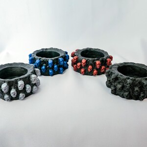 Pode incluir: Quatro castiçais circulares com base preta, cada um decorado com pequenas caveiras de cores diferentes. Um apresenta caveiras prateadas, um azuis, um vermelhas e um pretas. Os castiçais têm uma aparência texturizada e um centro oco.