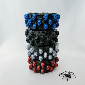 Pode incluir: Uma pilha de quatro recipientes cilíndricos com superfícies pretas texturizadas, cada um adornado com uma faixa de pequenas caveiras. O recipiente superior apresenta caveiras azul metálico, o segundo caveiras pretas, o terceiro prateadas e o inferior vermelhas.