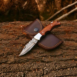 Peut inclure: Un couteau de poche pliant avec une lame en métal à motifs et un manche en bois poli, doté de rivets argentés. Le couteau est placé partiellement dans un étui en cuir marron foncé avec des coutures orange visibles et un bouton-pression en métal.
