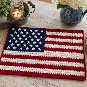 May include: A rectangular crochet placemat featuring the design of the American flag. The piece displays a dark blue field with white stars in the upper left corner, followed by alternating horizontal stripes in red and white across the surface.