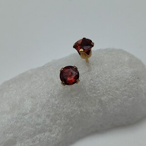 May include: A pair of stud earrings featuring round, deep red gemstones set in gold-colored metal. The earrings are displayed on a white, textured surface, showcasing their vibrant color and simple design. The gemstones appear to be faceted, catching the light.