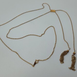 Puede incluir: Un collar lariat de color dorado con una delicada cadena y detalles de borlas. El collar presenta una barra deslizante para ajustar la longitud y dos borlas colgantes. El cierre es un peque&ntilde;o gancho dorado.