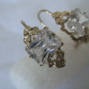 May include: A pair of gold earrings with clear, square-shaped gemstones. The earrings have a delicate, intricate design.