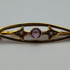 May include: A gold-toned brooch with an elongated, openwork design. It features a central, round, purple gemstone, flanked by two star-shaped accents, each with a small, round, white pearl. The brooch is a vintage jewelry piece.