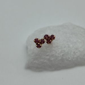 May include: A pair of gold stud earrings, each with three small, round, deep red gemstones in a cluster. The earrings are set against a soft, white background, accentuating their colour and design.
