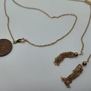 Puede incluir: Un collar dorado con una cadena y dos colgantes de borlas. Se coloca una moneda junto al collar para comparar el tama&ntilde;o. El collar tiene un cierre y una peque&ntilde;a cuenta redonda cerca de cada borla.