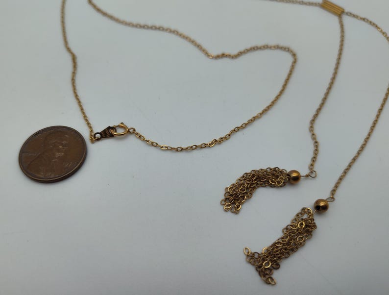 Puede incluir: Un collar dorado con una cadena y dos colgantes de borlas. Se coloca una moneda junto al collar para comparar el tama&ntilde;o. El collar tiene un cierre y una peque&ntilde;a cuenta redonda cerca de cada borla.