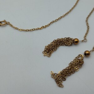Puede incluir: Un collar dorado con una cadena delicada y dos colgantes de borlas. Las borlas est&aacute;n hechas de m&uacute;ltiples hebras de cadena dorada, cada una acentuada con una peque&ntilde;a cuenta redonda. El collar se muestra sobre un fondo blanco.