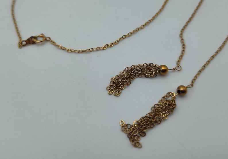Puede incluir: Un collar dorado con una cadena delicada y dos colgantes de borlas. Las borlas est&aacute;n hechas de m&uacute;ltiples hebras de cadena dorada, cada una acentuada con una peque&ntilde;a cuenta redonda. El collar se muestra sobre un fondo blanco.
