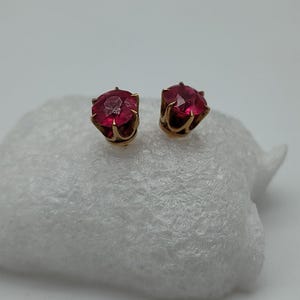 May include: A pair of stud earrings with round, faceted, ruby-red gemstones set in gold-coloured metal. The earrings are displayed on a white, textured surface against a plain background.
