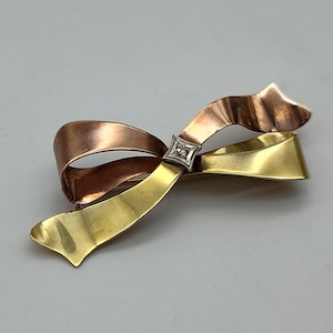 May include: A vintage bow brooch with a two-tone design. The top loop is copper-coloured, the bottom loop is gold-coloured, and the centre features a small, square, diamond-studded accent. The brooch is a classic jewellery piece.