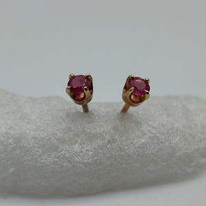 May include: A pair of gold stud earrings, each featuring a round, vibrant red gemstone. The earrings are set in a classic four-prong setting, showcasing the gemstones against a neutral background.