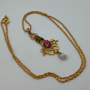 May include: A gold-colored necklace with a pendant featuring a red gemstone and a pear-shaped pearl drop. The pendant has an ornate, filigree design with leaf accents. The chain is a delicate, twisted rope style.