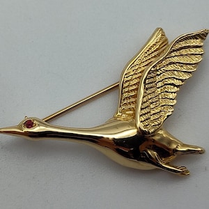 May include: A gold-colored brooch in the shape of a flying goose. The goose has detailed wings and a red gemstone eye. The brooch is made of a shiny, reflective metal and is set against a plain background.