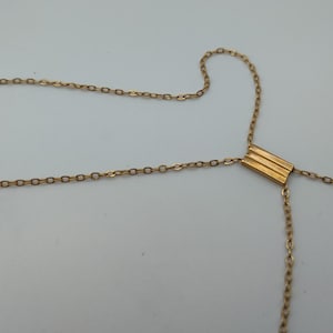 Puede incluir: Collar dorado con un dise&ntilde;o de cadena delicado. El collar presenta un colgante rectangular con tres l&iacute;neas horizontales. La cadena se extiende en forma de Y, creando un aspecto &uacute;nico y elegante.