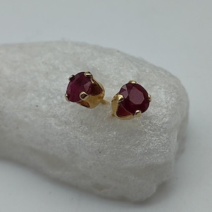 Peut inclure: Une paire de boucles d'oreilles à clous avec des pierres précieuses rondes, rouge foncé, serties dans du métal doré. Les boucles d'oreilles sont présentées sur une surface blanche et texturée, mettant en valeur leur couleur et leur design. La monture dorée met en valeur la couleur riche des pierres.