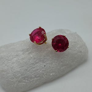 May include: A pair of gold-colored stud earrings, each featuring a large, round, deep red gemstone. The earrings are set on a white, textured surface, creating a contrast that highlights the vibrant color of the stones. The earrings are a classic jewelry item.