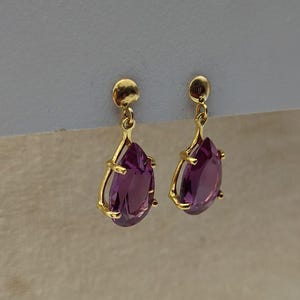 May include: A pair of gold-colored drop earrings featuring pear-shaped, deep purple gemstones. The earrings have a post-and-stud design with a decorative gold frame around the gemstones. The earrings are displayed against a neutral background.