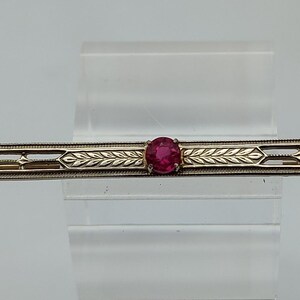 May include: A long, slender bar brooch crafted from a metallic material with a warm, golden tone. The brooch features an elongated, openwork design with engraved leaf motifs and a single, round, vibrant pink gemstone set in the center.