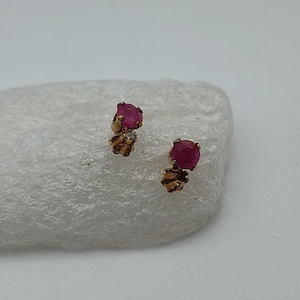 May include: A pair of gold-colored stud earrings, each featuring a round, deep red gemstone. The earrings are set on a textured, organic-shaped base, creating a classic and elegant look. The gemstones are approximately 0.25 inches in diameter.