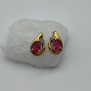 May include: A pair of gold-coloured stud earrings. Each earring has a teardrop-shaped, vibrant red gemstone, with small, clear stones. The earrings are displayed on a white, textured surface.