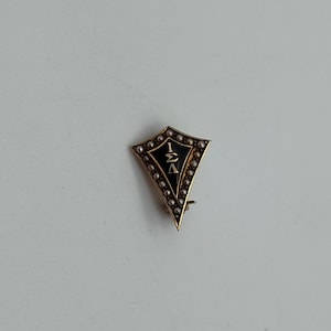 May include: A gold-toned, shield-shaped pin with a black center. The center features white Greek letters and is surrounded by small, pearl-like beads. The pin has a clasp on the back.