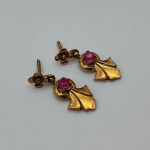 May include: A pair of gold-coloured earrings. Each earring has a floral stud and a dangling element. They feature a round, red gemstone set in a gold frame, with a leaf shape below. The earrings are shown against a white background.