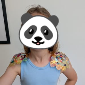 Puede incluir: Una camiseta sin mangas de color azul claro con hombreras decorativas adornadas con una variedad de lentejuelas iridiscentes multicolores y adornos en forma de flor que crean una superficie texturizada y reflectante en ambos hombros.