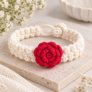 May include: A cream-colored crocheted bracelet featuring a textured, knotted band and a prominent, vibrant red crocheted rose at the center. The bracelet includes a circular loop and a matching crocheted button closure for a secure fit.