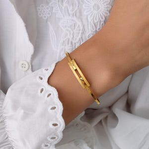 Puede incluir: Un brazalete de metal en tono dorado que presenta un diseño de barra rectangular central con un centro hueco. El brazalete tiene un acabado liso y pulido, con una banda circular delgada que rodea la muñeca, mostrando una estética limpia y geométrica.
