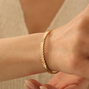 Pode incluir: Uma pulseira de tom dourado com um padrão repetitivo de chevron ou em forma de seta. Os elos metálicos estão dispostos numa sequência angular contínua que envolve o pulso, criando uma aparência geométrica texturizada com um acabamento polido.