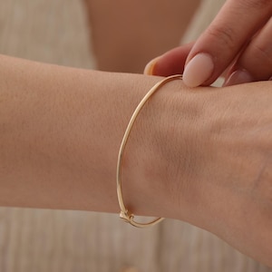 Puede incluir: Una pulsera tipo brazalete de oro pulido, delgada, con una superficie lisa y redondeada y un pequeño mecanismo de cierre de bisagra integrado. La pulsera presenta un diseño circular sencillo que se ajusta cómodamente a la muñeca, mostrando un acabado metálico brillante.