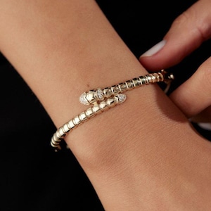 Könnte beinhalten: Ein goldenes Armband in Schlangenform mit einem segmentierten Körperdesign, das sich um das Handgelenk legt. Der Schlangenkopf ist mit kleinen, klaren Pavé-Steinen verziert, die einen schimmernden Effekt auf der Metalloberfläche des Schmuckstücks erzeugen.