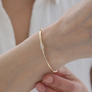 Puede incluir: Una pulsera rígida de oro pulido y fino que presenta una pieza central rectangular engastada con varias piedras pequeñas y claras de corte baguette. La banda de metal tiene un acabado liso y reflectante con un cierre visible.