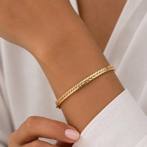 Könnte beinhalten: Ein goldfarbenes Armband mit einem flachen, gewebten Fischgrätenmuster. Das Armband hat eine polierte metallische Oberfläche und enthält einen kleinen, integrierten Verschlussmechanismus, der an der Seite des Handgelenks sichtbar ist.
