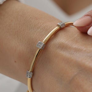 Op de afbeelding: Een goudkleurige armband met drie gelijkmatig verdeelde, zilverkleurige vierkante elementen. Elk vierkant element is versierd met kleine, heldere, fonkelende steentjes in een rasterpatroon op de gladde, gepolijste goudkleurige metalen band.