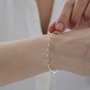 May include: A gold bangle bracelet featuring a scalloped edge design. The bracelet is set with small, round, vibrant pink gemstones alternating with tiny, clear, sparkling white stones along the entire outer curved surface of the metal band.