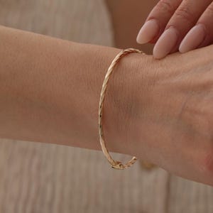 Könnte beinhalten: Ein dünner, goldfarbener Armreif mit einer gedrehten, seilartigen Textur entlang des gesamten Bandes. Der kreisförmige Armreif verfügt über einen kleinen, integrierten Scharniermechanismus zum Öffnen und Schließen sowie eine polierte Oberfläche.