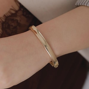 Op de afbeelding: Een goudkleurige visgraatkettingarmband met een plat, flexibel en in elkaar grijpend schakelontwerp. De armband heeft een gepolijst, reflecterend oppervlak dat het licht opvangt, wat zorgt voor een gladde, doorlopende uitstraling om de pols.