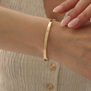 Könnte beinhalten: Ein polierter, goldfarbener Scharnierarmreif mit glatter, flacher Oberfläche und einem sicheren Verschlussmechanismus. Das Armband zeichnet sich durch ein schlichtes Design mit gleichmäßiger Breite aus und umschließt das Handgelenk mit einem glänzenden, metallischen Finish.