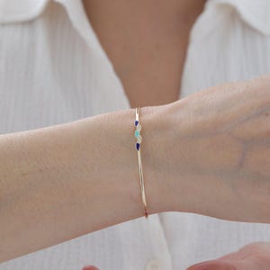 Könnte beinhalten: Ein dünner goldener Armreif mit einem zentralen Dekorelement, das aus zwei kleinen dunkelblauen tropfenförmigen Akzenten, einem hellblauen tropfenförmigen Akzent und zwei winzigen klaren Steinen in einer vertikalen Linie besteht.