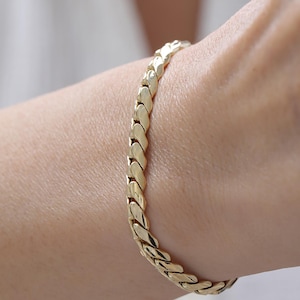 Könnte beinhalten: Ein goldfarbenes Kettenarmband mit einer Reihe von ineinandergreifenden, polierten, geschwungenen Gliedern, die eine durchgehende, strukturierte und reflektierende Oberfläche bilden. Das Armband liegt am Handgelenk und zeigt ein gleichmäßiges, sich wiederholendes Muster aus glatten Metallsegmenten.