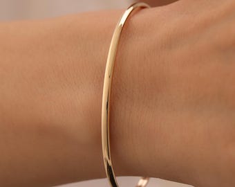 Puede incluir: Una pulsera tipo brazalete de oro pulido con una superficie lisa y redondeada y un mecanismo de cierre de bisagra seguro. La pulsera presenta un diseño circular continuo y elegante que mantiene un grosor constante en toda su circunferencia.