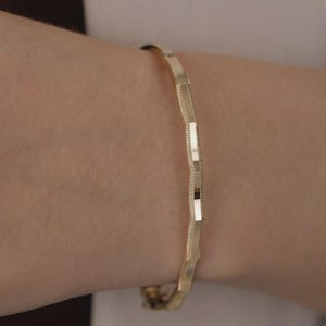 Op de afbeelding: Een gouden armband bestaande uit een reeks verbonden, gefacetteerde segmenten. Elk segment wisselt af tussen een glad, gepolijst oppervlak en een fijn getextureerde, geribbelde afwerking, wat een geometrisch patroon rond de pols vormt.